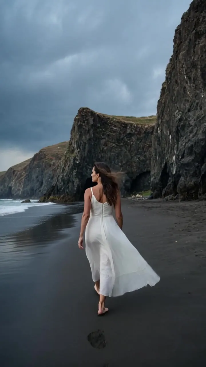 A woman in a white dress walking a black sand beach under storm clouds
